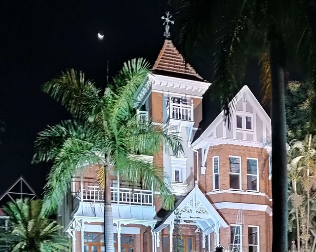 Illuminated Victorian house with a tower and palm trees beneath a crescent moon